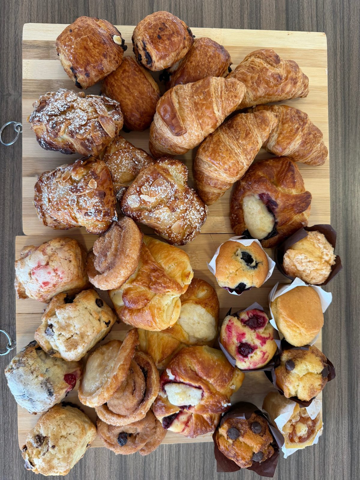 An assortment of mini croissants, almond croissants, danishes, scones and muffins on a wooden board